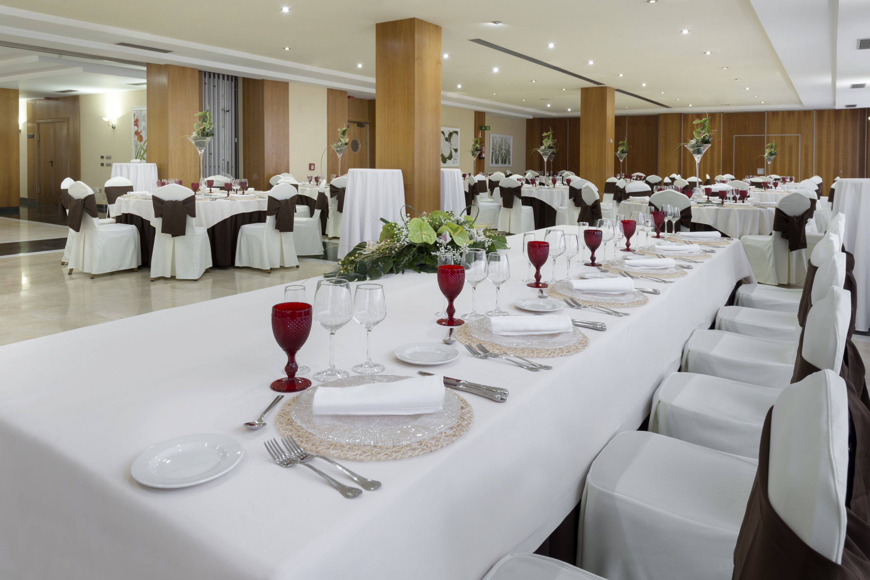 A banquet hall with round tables set for a formal event, complete with white tablecloths, red wine glasses, and silverware.