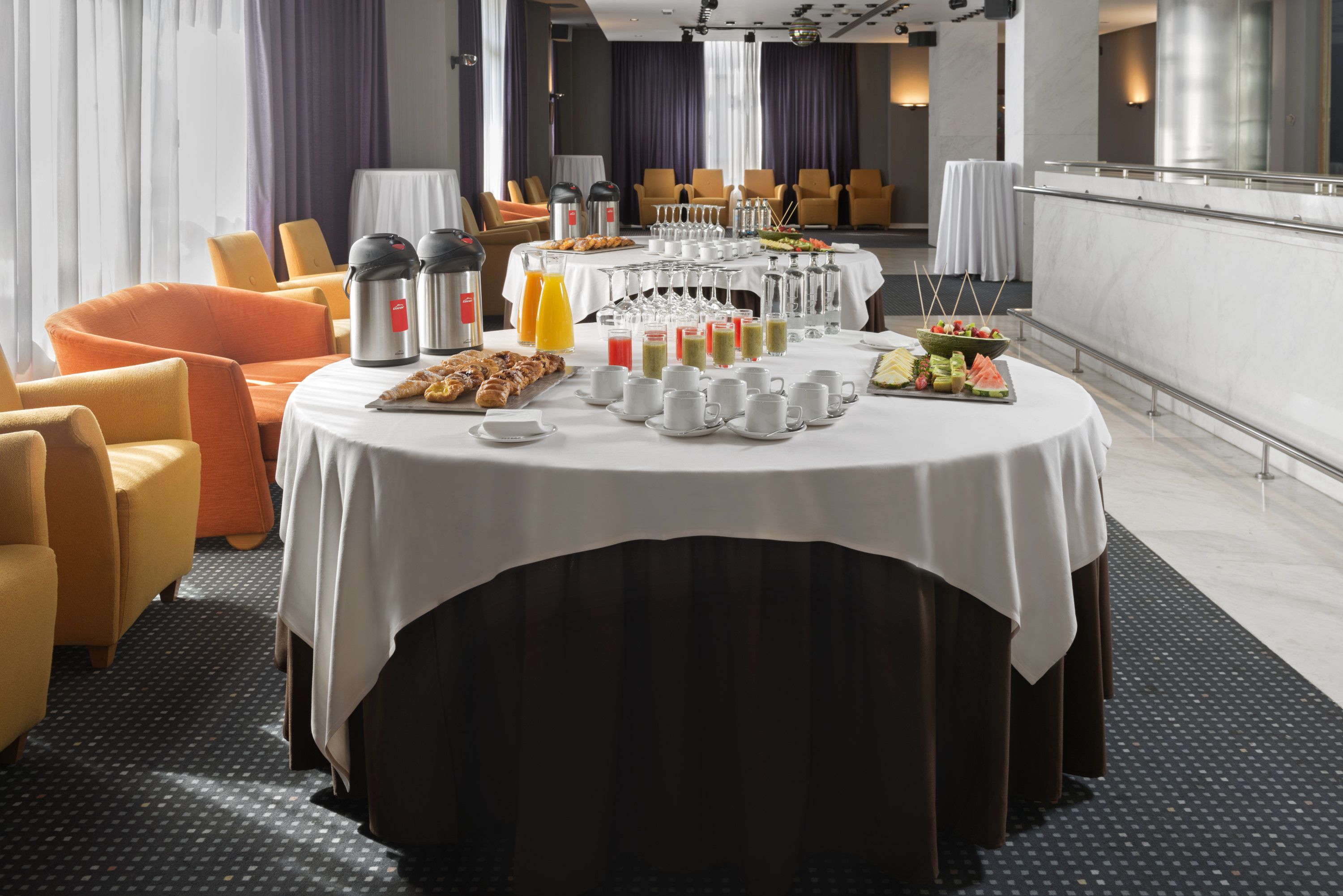 A breakfast buffet setup with various food items and beverages on a round table, surrounded by comfortable seating.