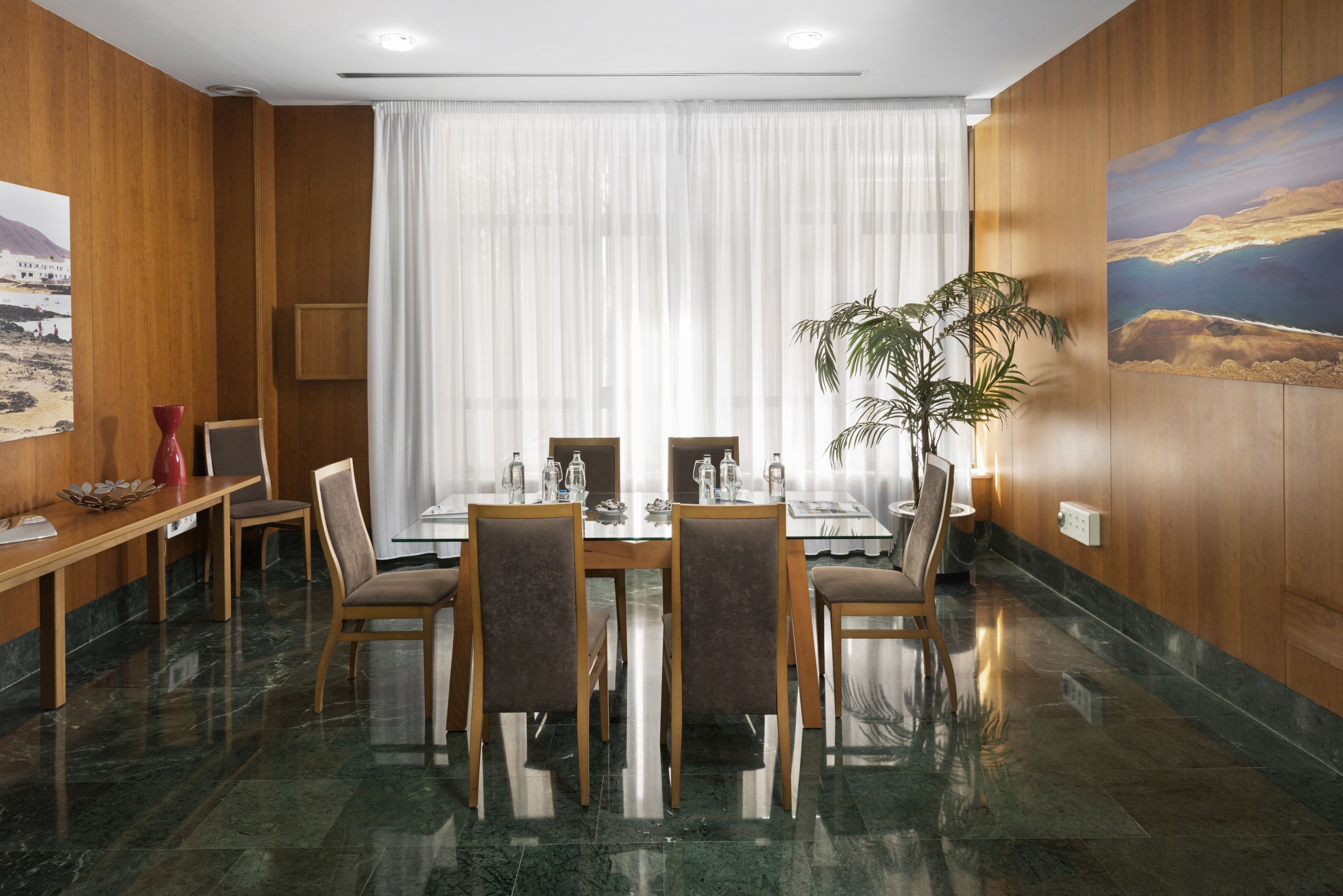 A hotel dining area with a wooden table set for six, surrounded by chairs, a potted plant, and a large window with sheer curtains.