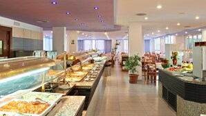 Un área de bufé con varias estaciones de comida, un espacio de comedor con mesas y sillas, y una planta en maceta.