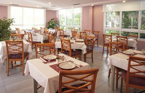 A dining area with multiple tables and chairs, large windows with natural light, and potted plants. - Club Hotel Cala Ratjada (Capdepera)