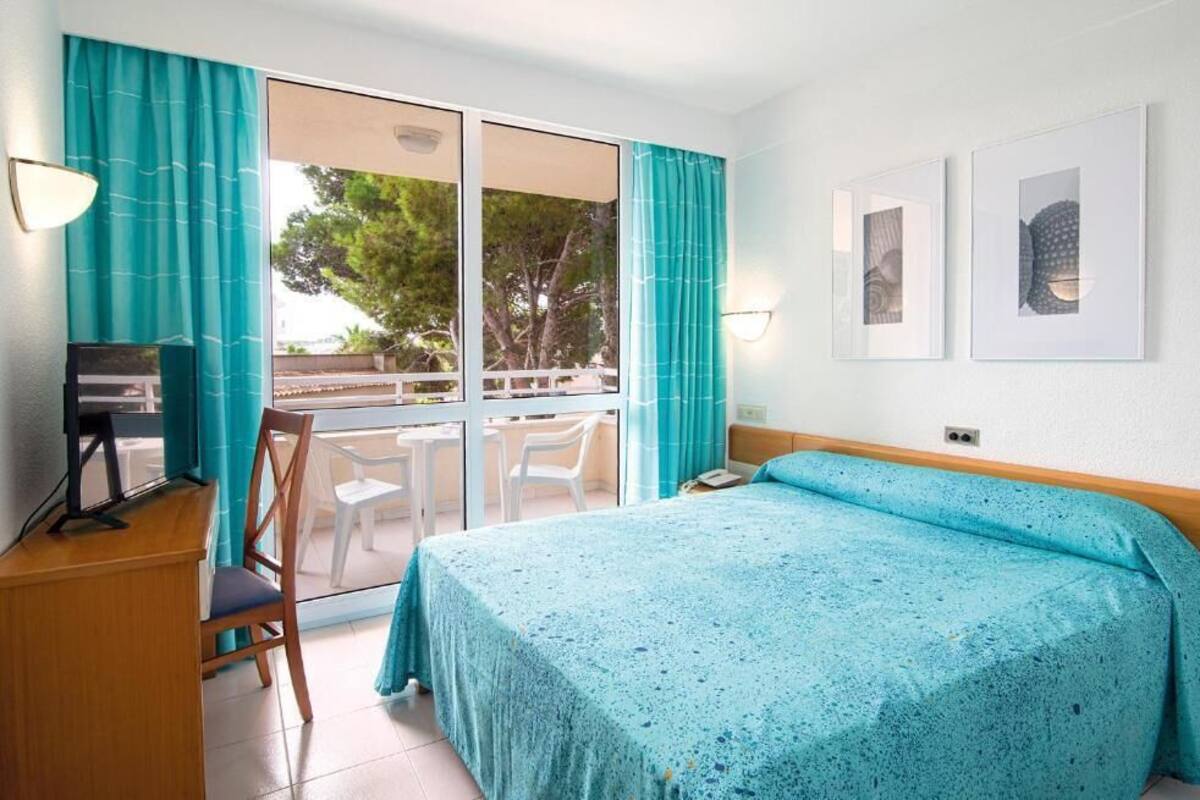 A bedroom with a bed, a desk, and a chair. There are two windows with curtains, and a balcony with chairs.