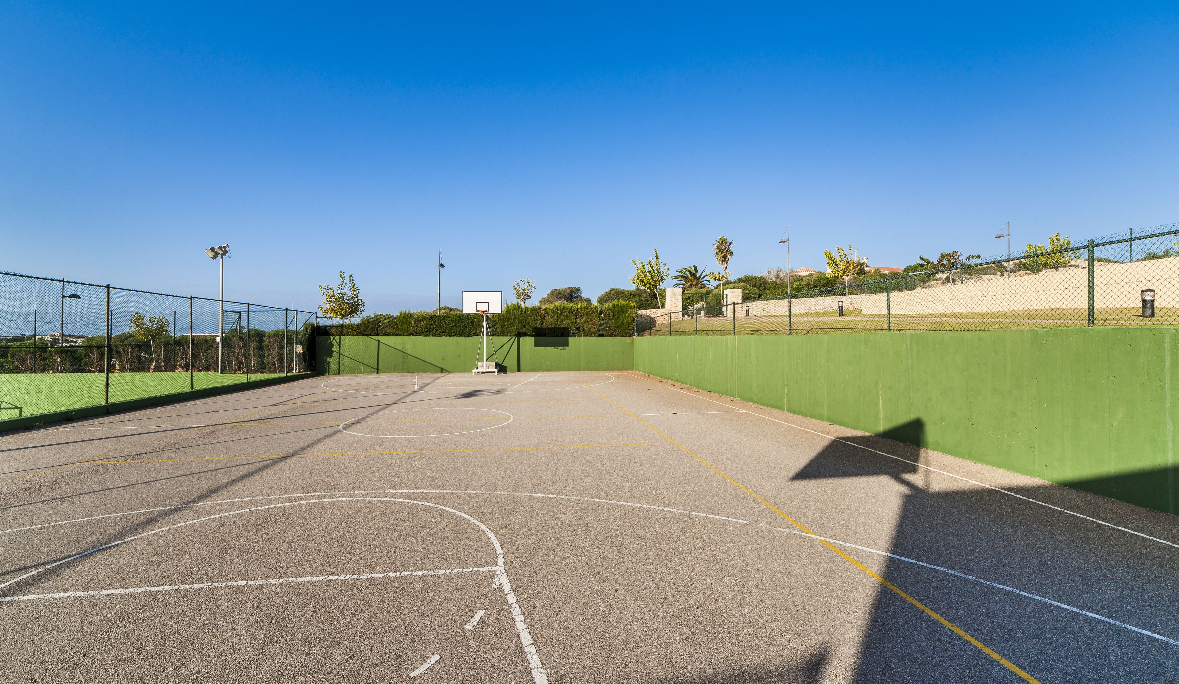 An outdoor basketball court with a green fence, clear sky, and distant buildings.