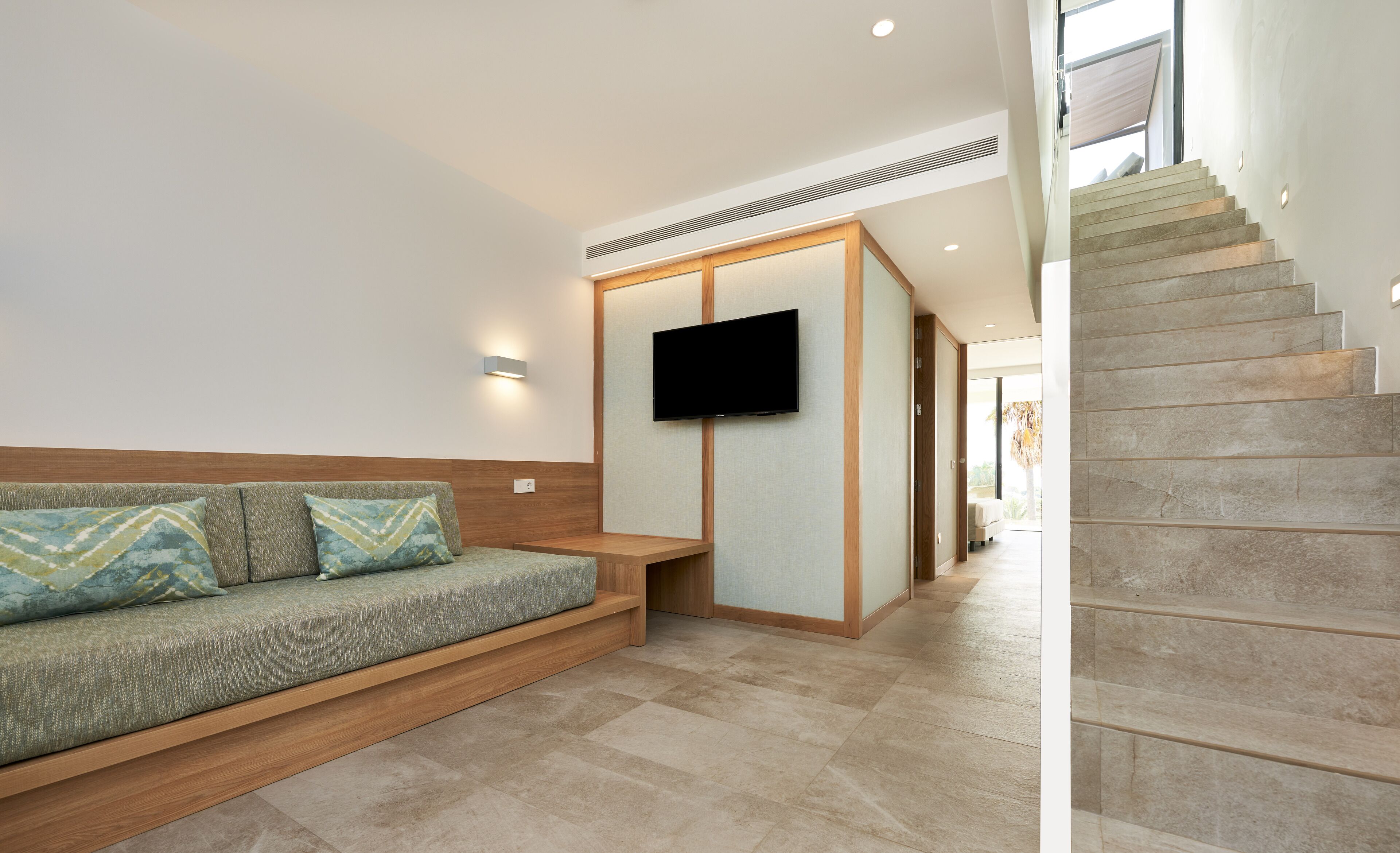 A modern living room with a sofa, a flat-screen TV mounted on a wall, and a staircase leading to an upper level.