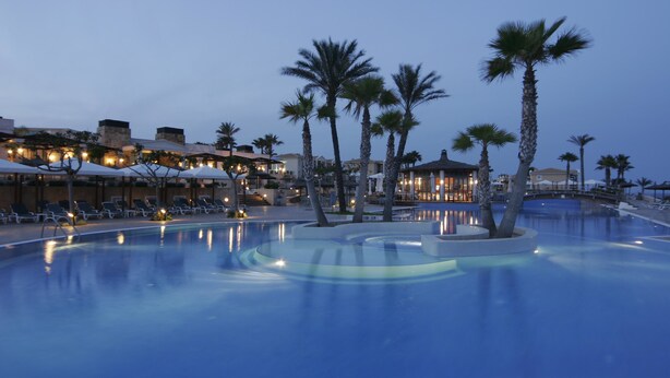 A pool area with lounge chairs and umbrellas, surrounded by palm trees and buildings with outdoor seating. - Insotel Punta Prima Resort & Spa (Sant Lluis)