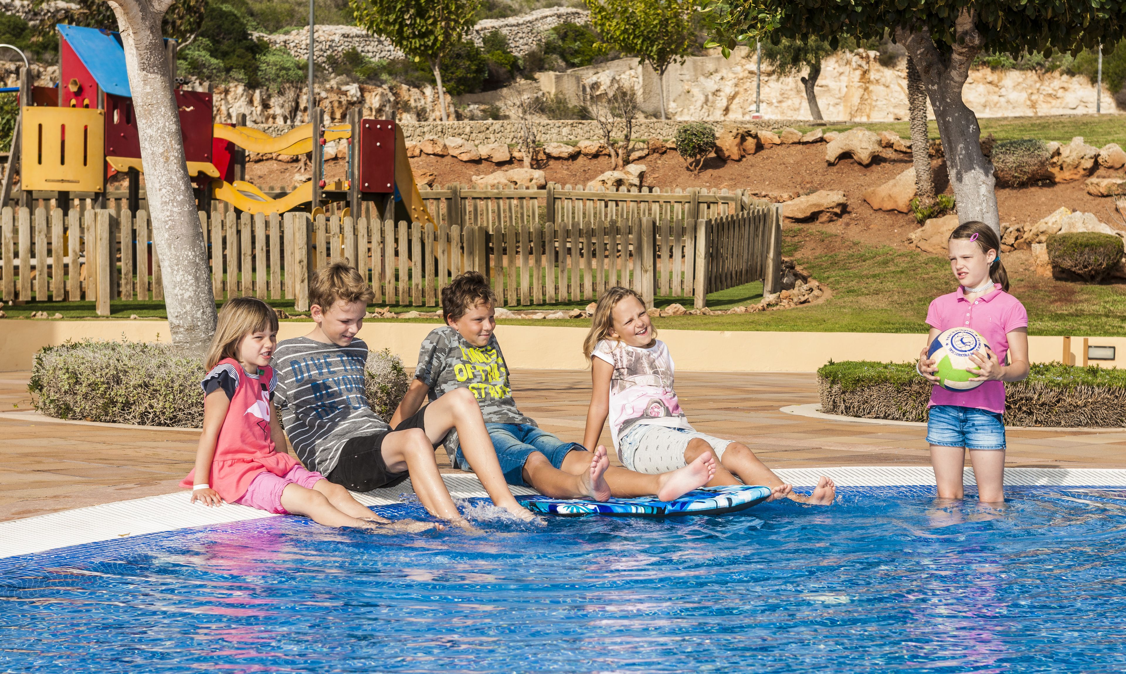 A group of children sitting by a pool with a wooden fence and playground in the background.