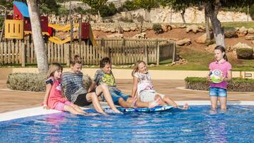 A group of children sitting by a pool with a wooden fence and playground in the background.