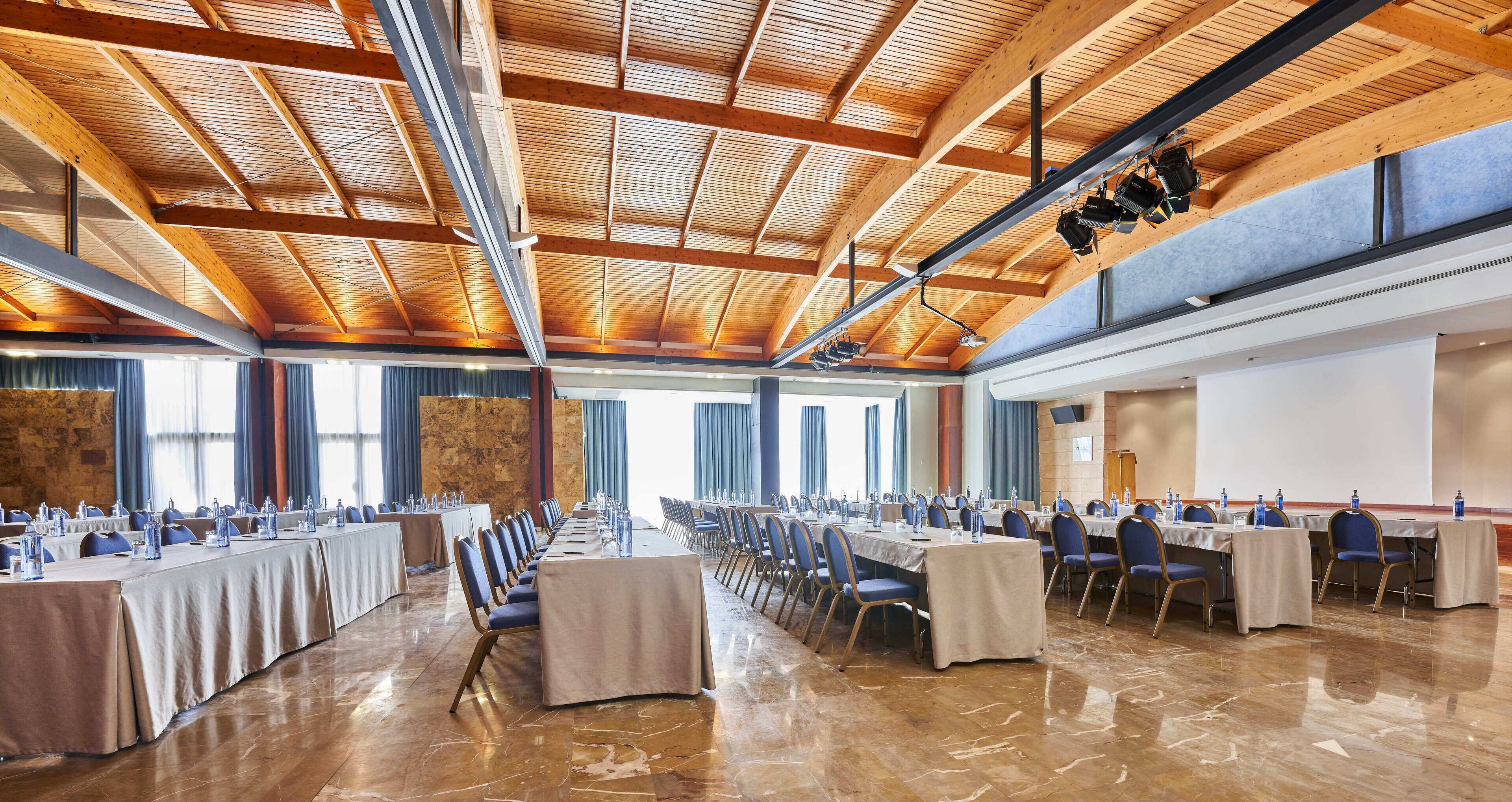 Un salón de conferencias con largas mesas, sillas y un alto techo de madera con vigas a la vista.