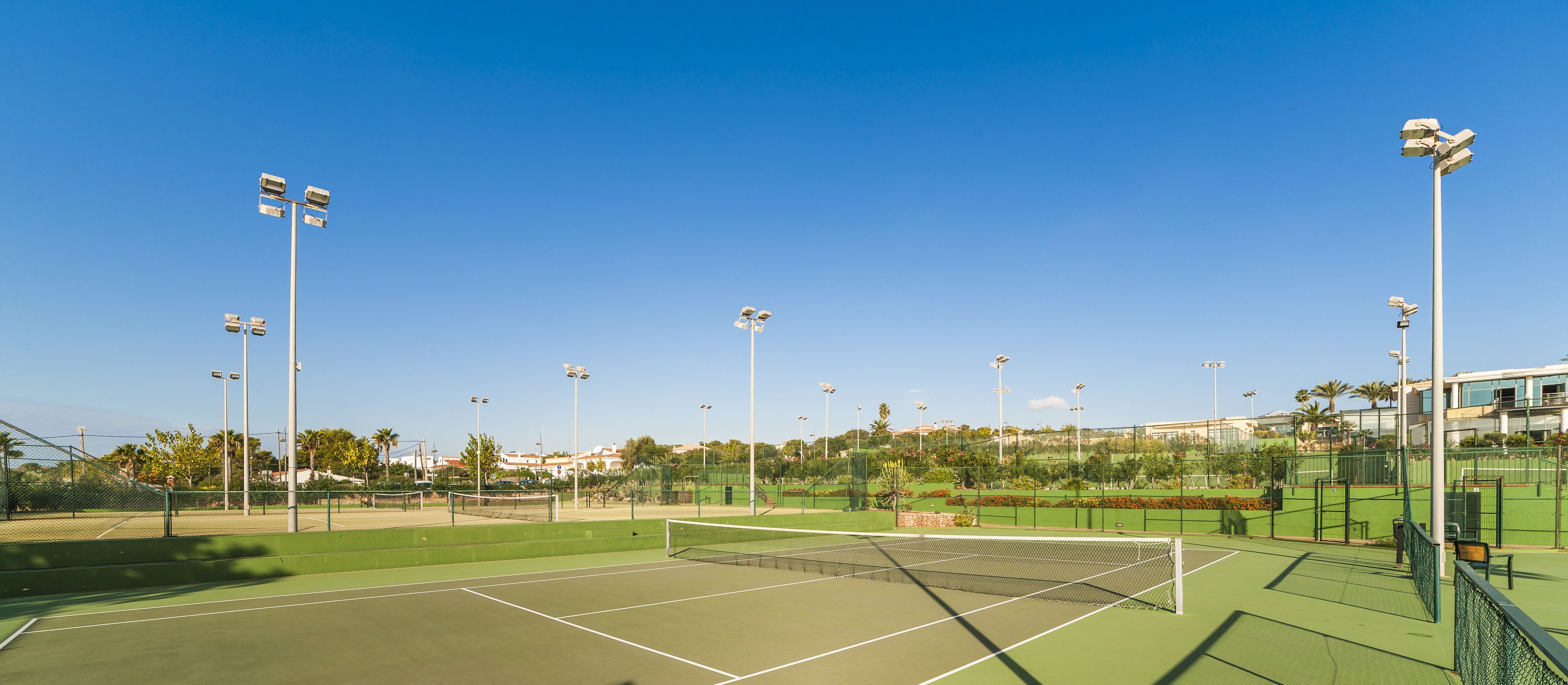 A tennis court with multiple light poles, surrounded by greenery and buildings in the background.