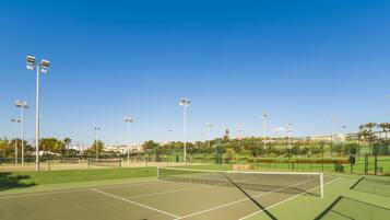 A tennis court with multiple light poles, surrounded by greenery and buildings in the background.