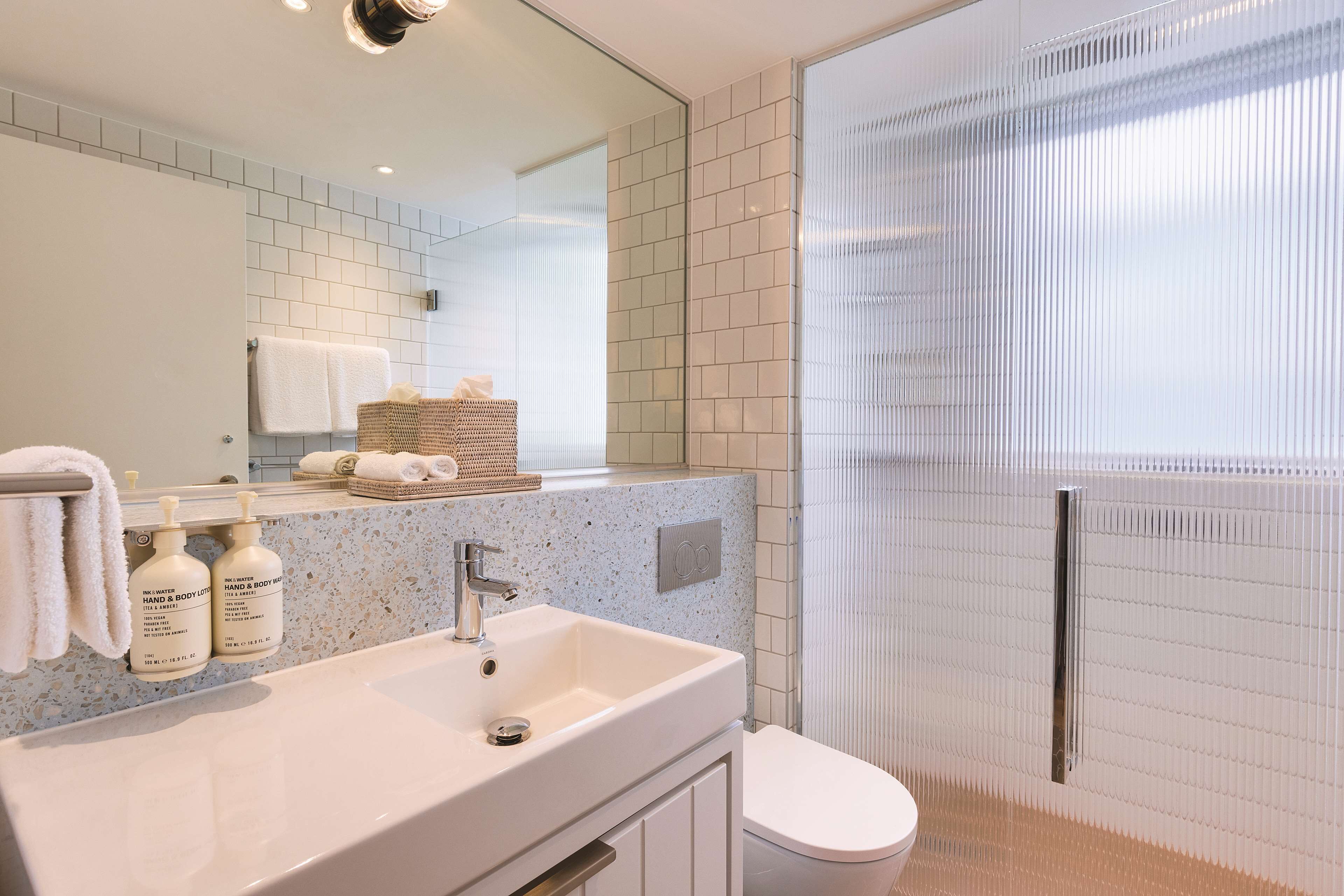 A modern bathroom with a white sink, two bottles of hand and body lotion, a towel rack, and a glass shower enclosure.