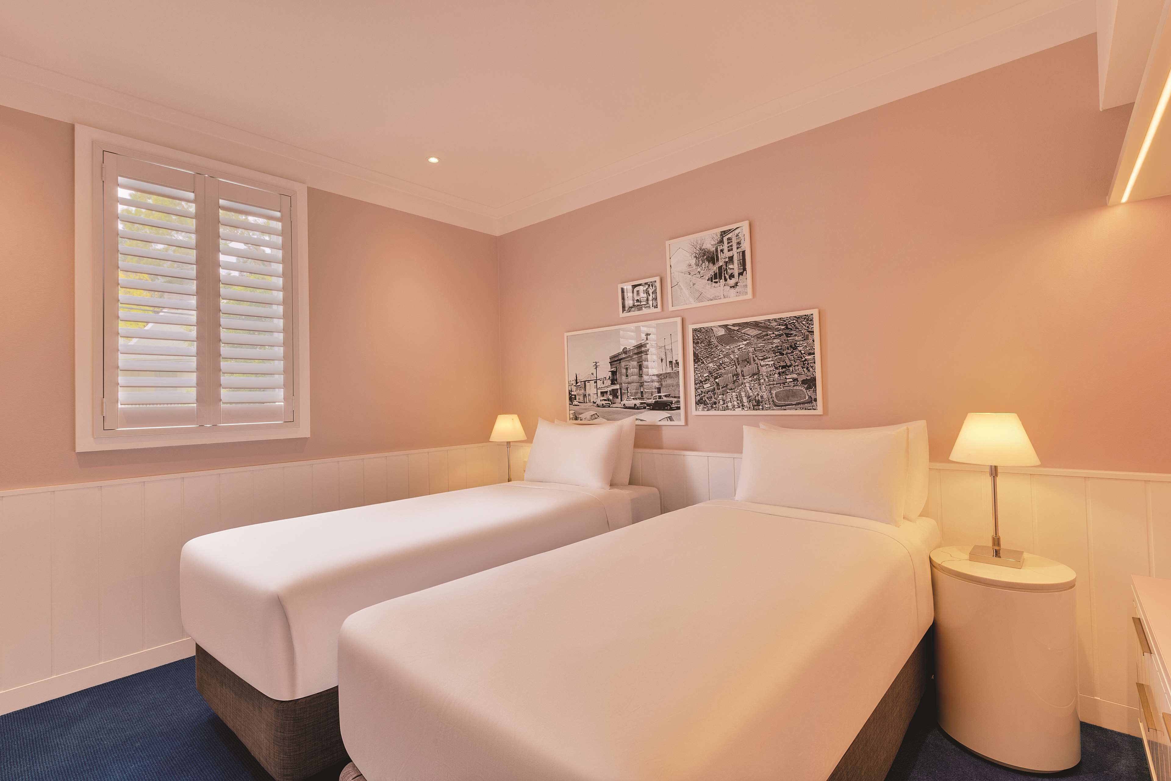 A hotel room with two beds, a window with blinds, a wall with framed pictures, and two bedside lamps.