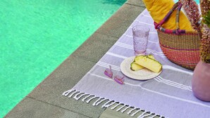 A poolside setting with a woven basket containing a yellow towel, a glass of water, and a plate with pineapple slices.