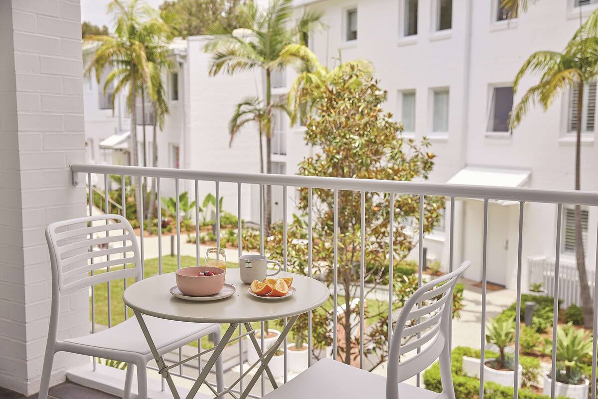Un balcon avec une table et des chaises blanches, un saladier rempli de fruits et une tasse.
