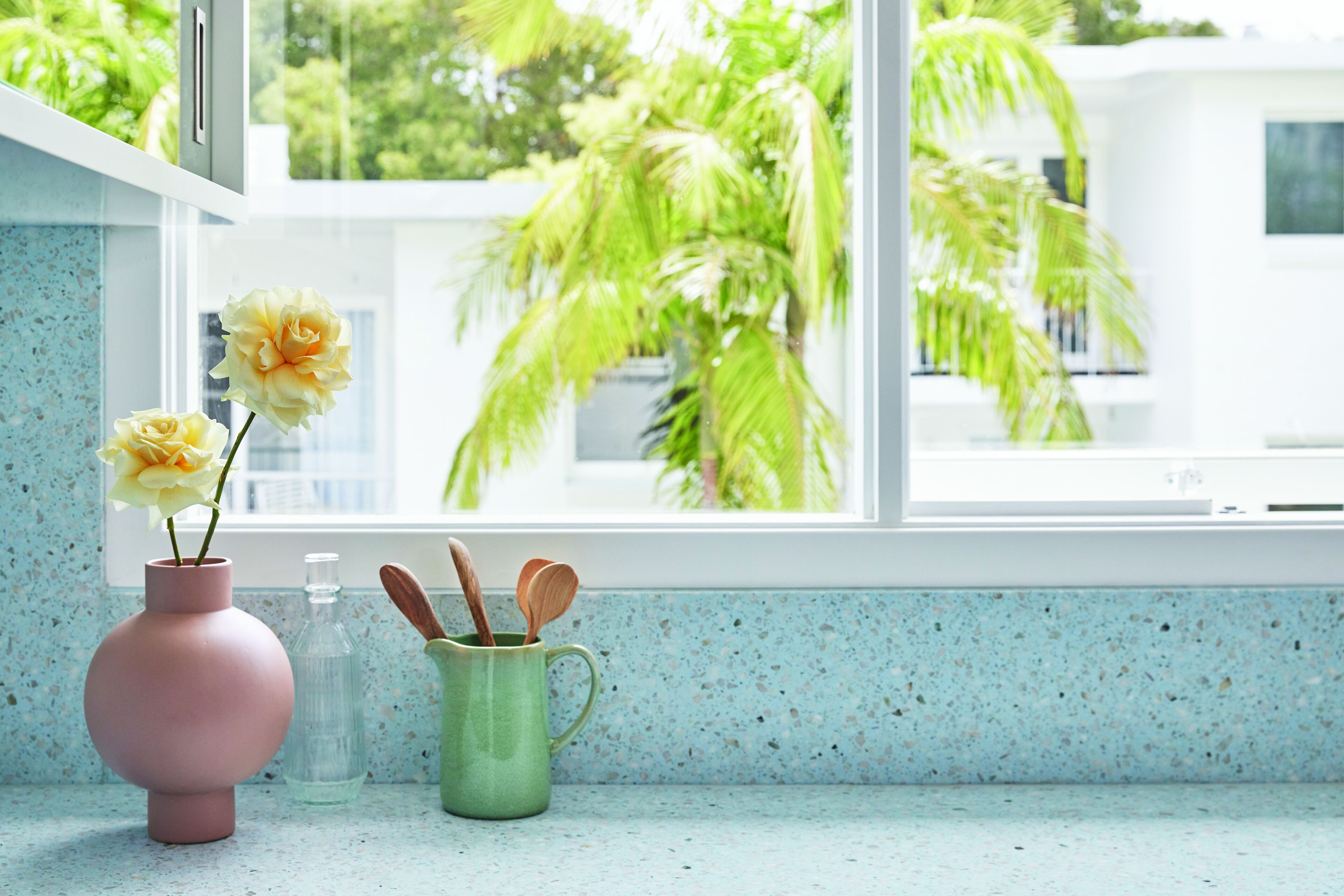 A kitchen counter with a pink vase, a green mug, and a clear bottle.