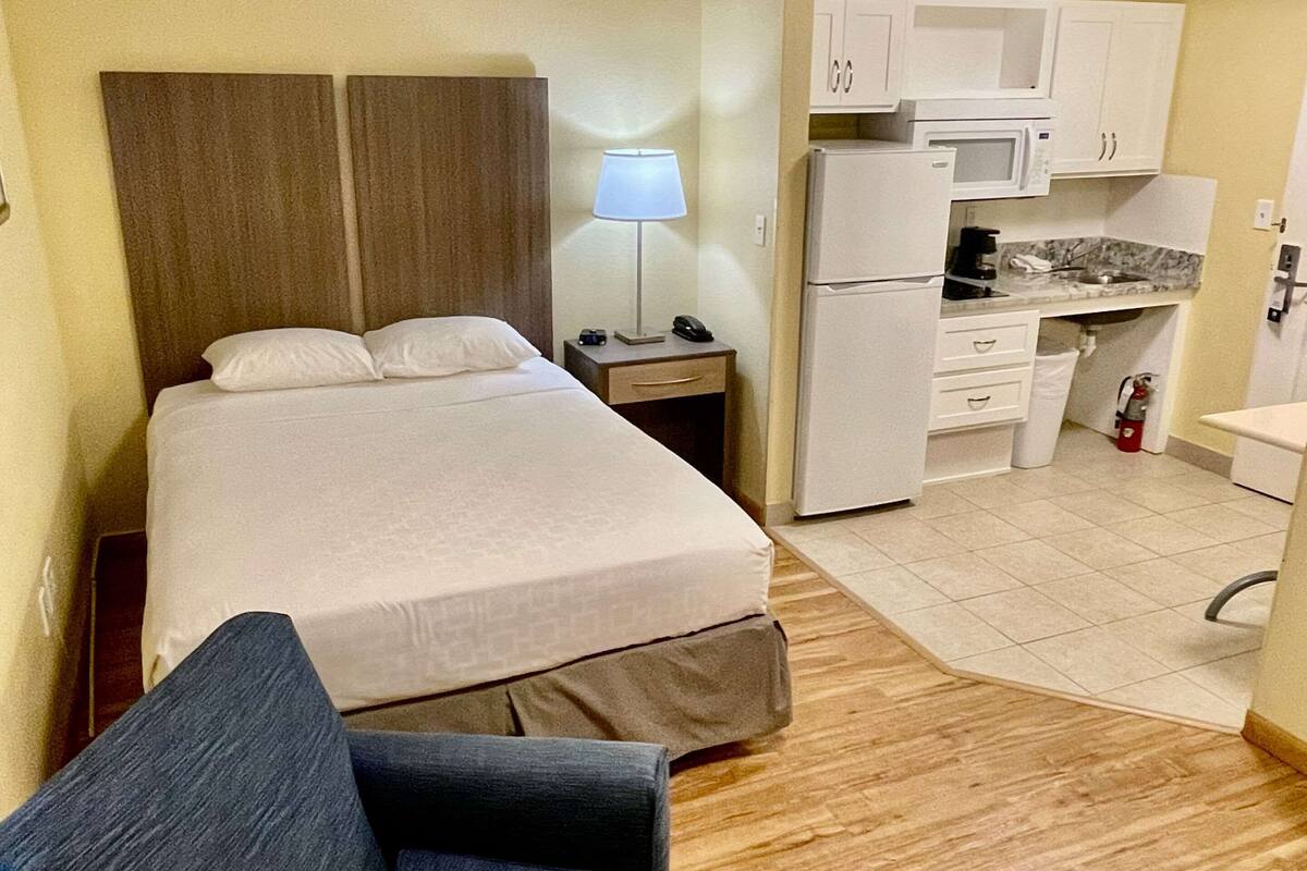 Ein Hotelzimmer mit Bett, Nachttisch mit Lampe, einer kleinen Küchenzeile inklusive Kühlschrank, Mikrowelle und Kaffeemaschine.