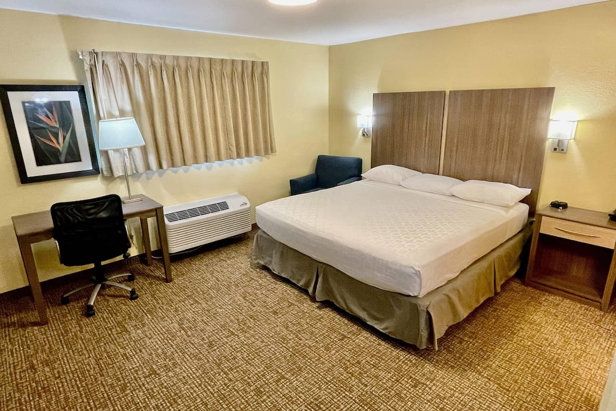 Ein Hotelzimmer mit einem Bett, einem Schreibtisch mit Stuhl, einer Lampe, einem Fenster mit Vorhängen und einer Wand-Klimaanlage.