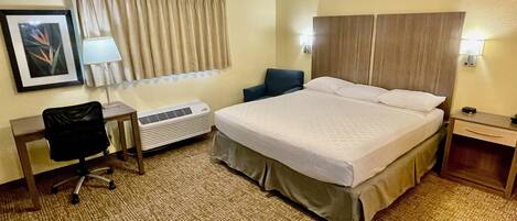 Een hotelkamer met een bed, een bureau met stoel, een lamp, een raam met gordijnen en een wandairconditioner.