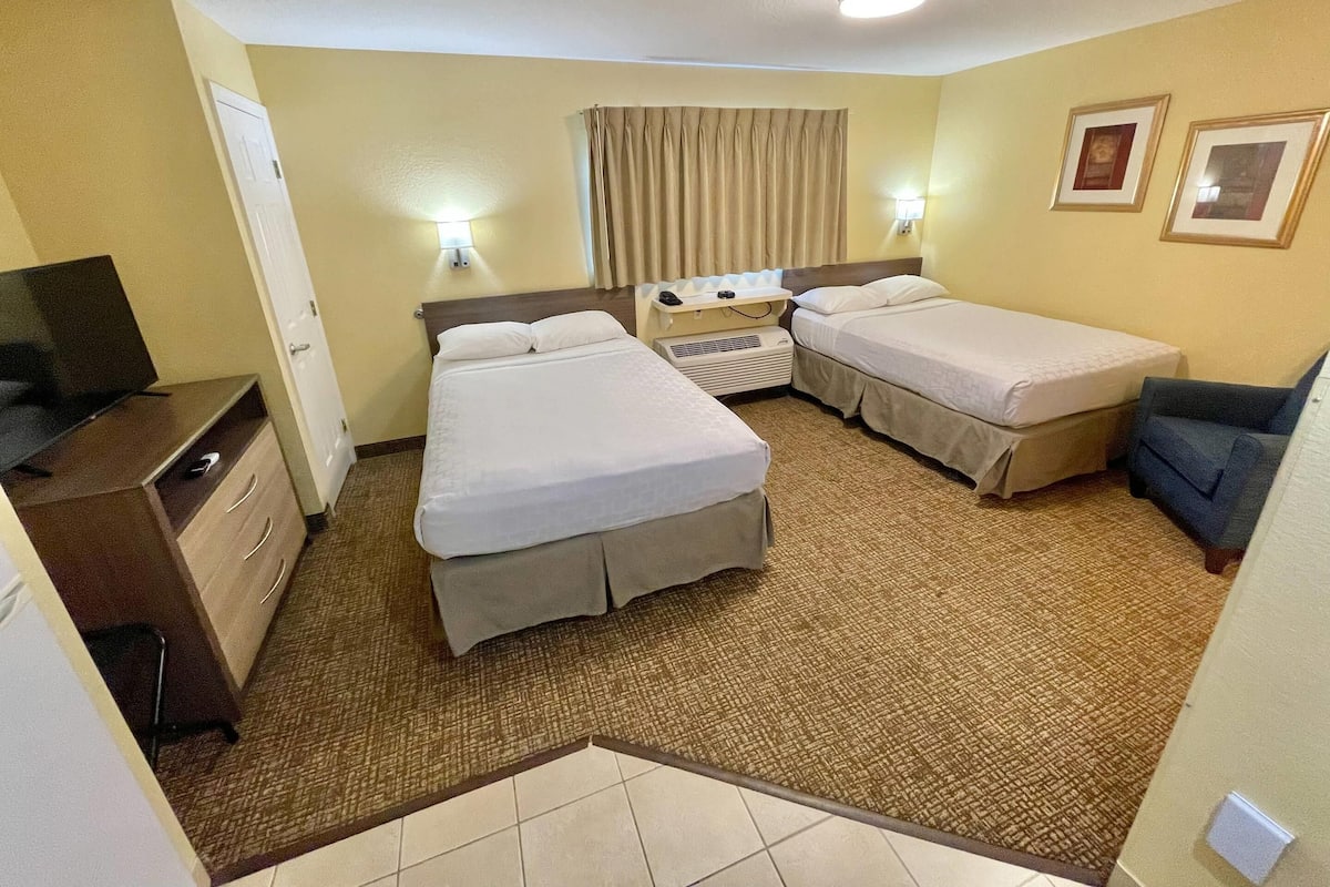 Ein Hotelzimmer mit zwei Betten, einem Schreibtisch mit Fernseher, einem Sessel und einem kleinen Tisch mit Lampe.