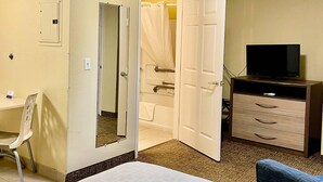 Ein Hotelzimmer mit einem Bett, einem Schreibtisch mit Stuhl, einem Fernseher auf einer Kommode und einem Badezimmer mit Duschvorhang.