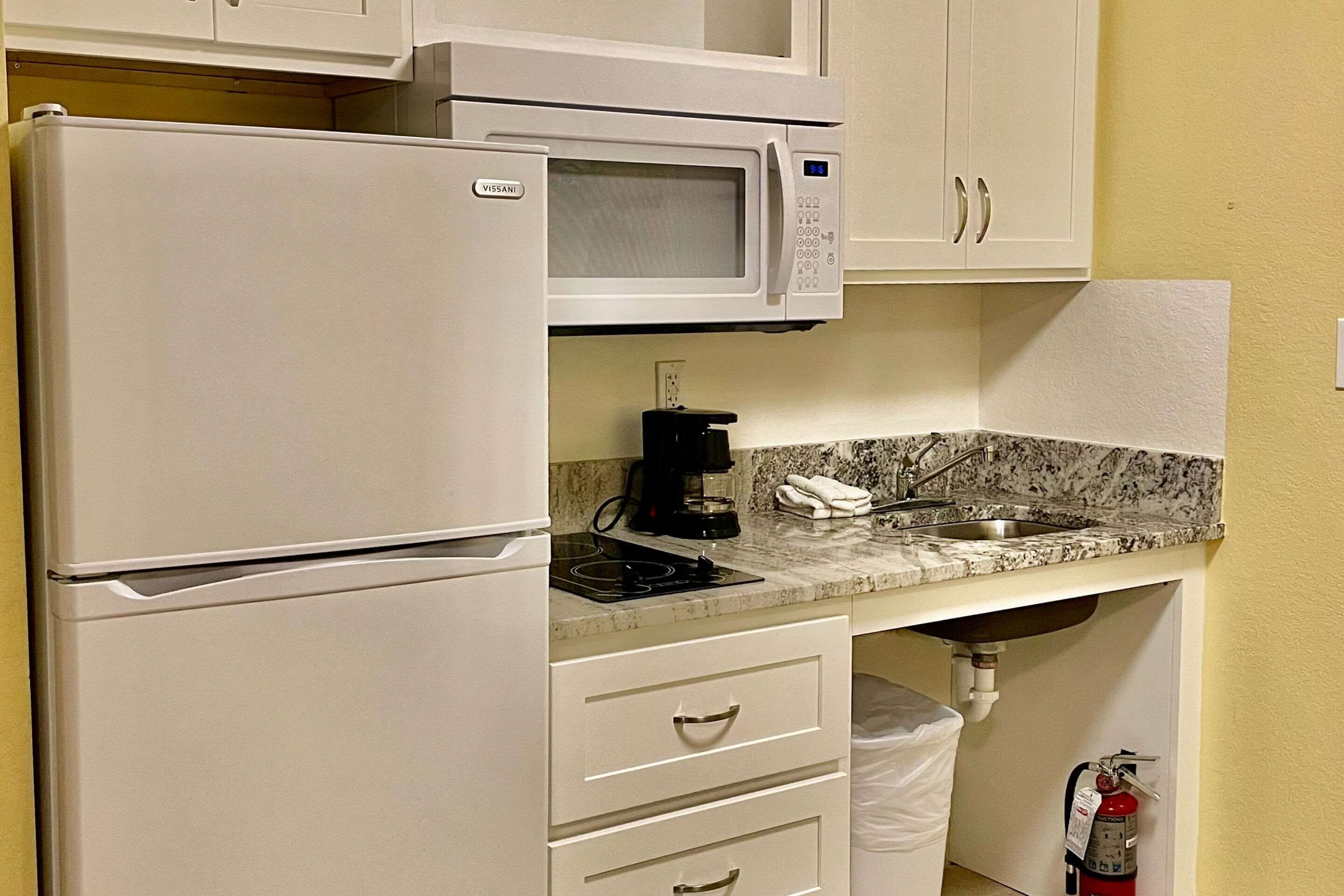 A kitchenette with a white refrigerator, microwave, and stove. There are cabinets above and below the counter, and a fire extinguisher is mounted on the wall.
