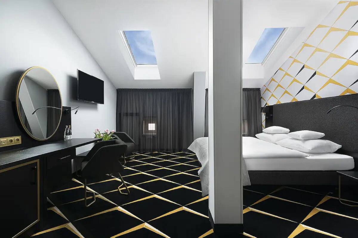 A modern hotel room with a black and yellow patterned floor, a large mirror, a flat-screen TV, a bed with white bedding, and a skylight above the bed.