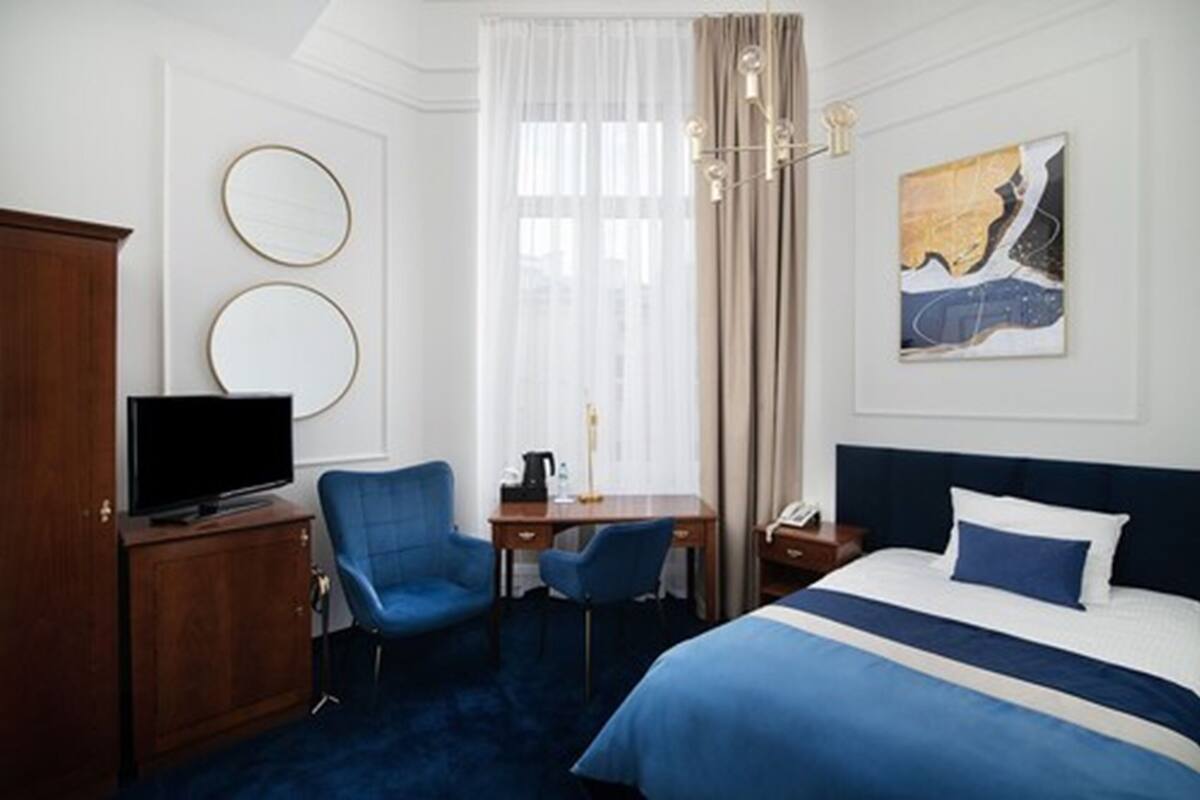 A hotel room with a bed, two chairs, a desk, a television, and a painting on the wall.