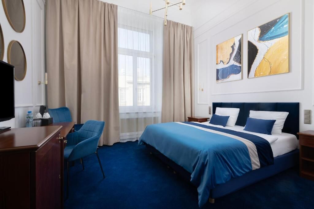 A hotel room with a bed, a desk, a chair, and artwork on the wall.