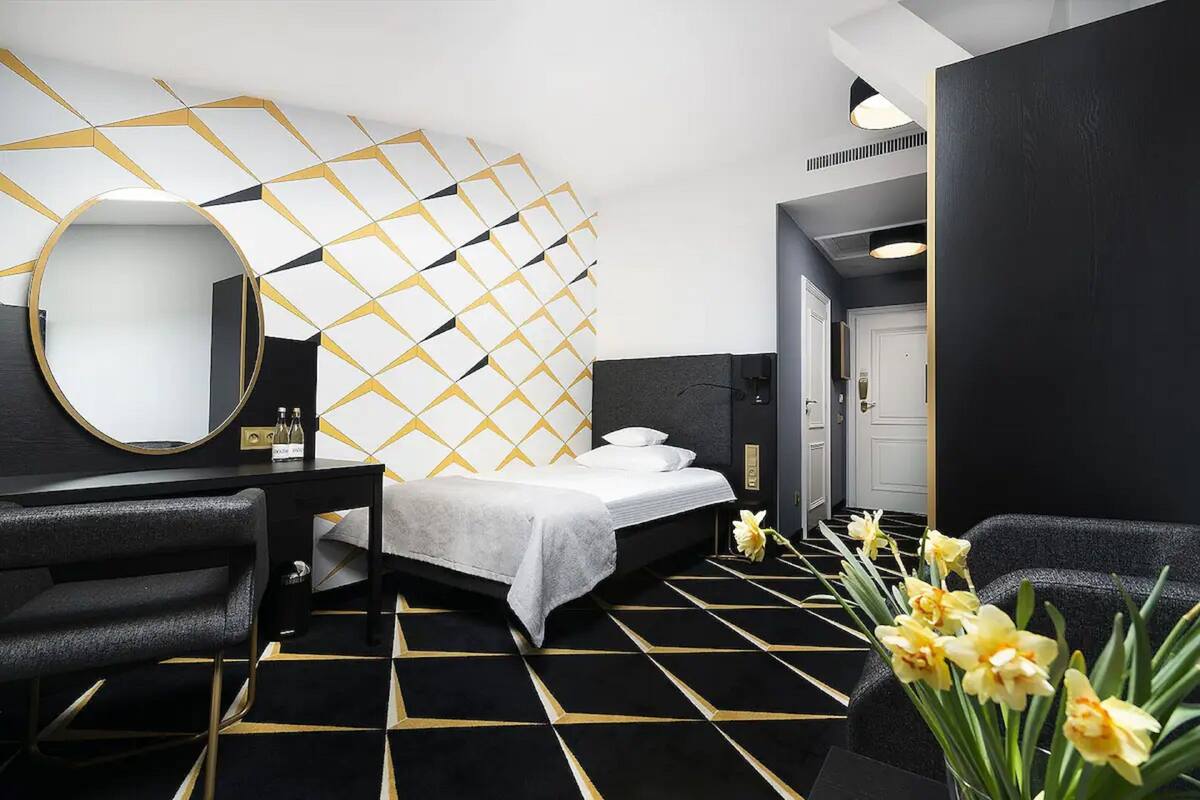 A modern hotel room with a geometric patterned wall, a round mirror, a bed with white linens, a black and gold floor, and a small plant with yellow flowers.