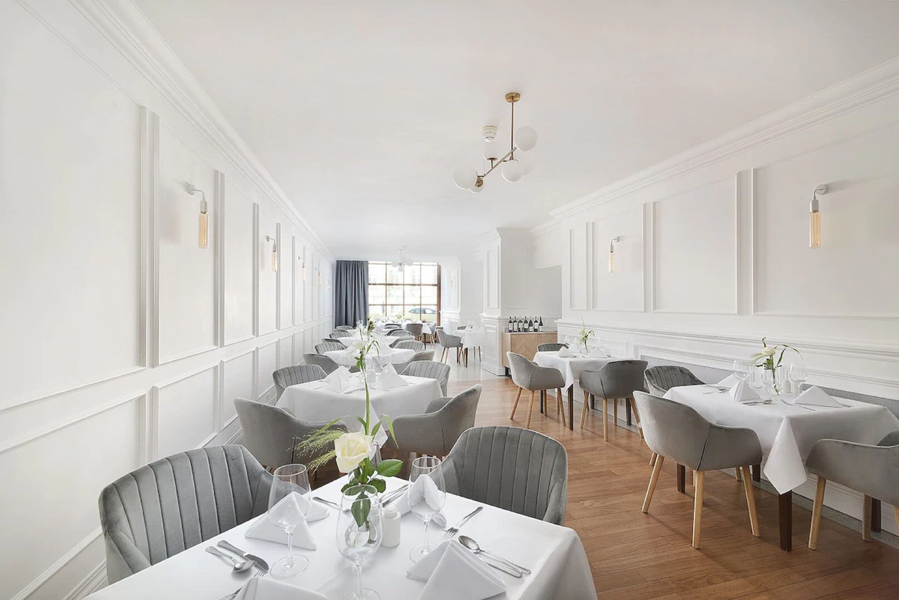 A dining area with multiple tables set for service, featuring white tablecloths and grey chairs.