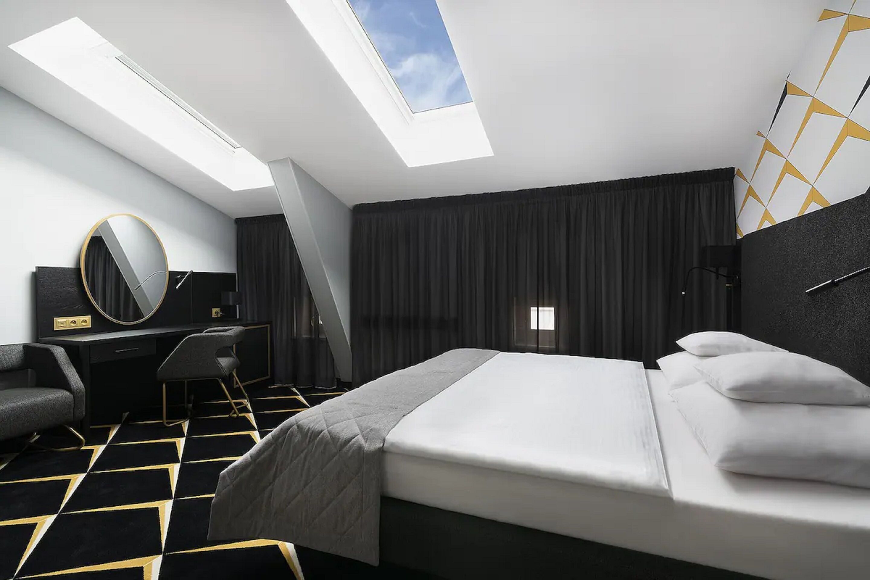 A modern hotel room with a skylight, a bed with white bedding, a desk with a chair, and a large mirror.