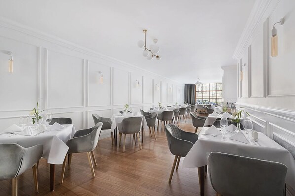 A spacious dining area with multiple tables and chairs, white walls, and a wooden floor.