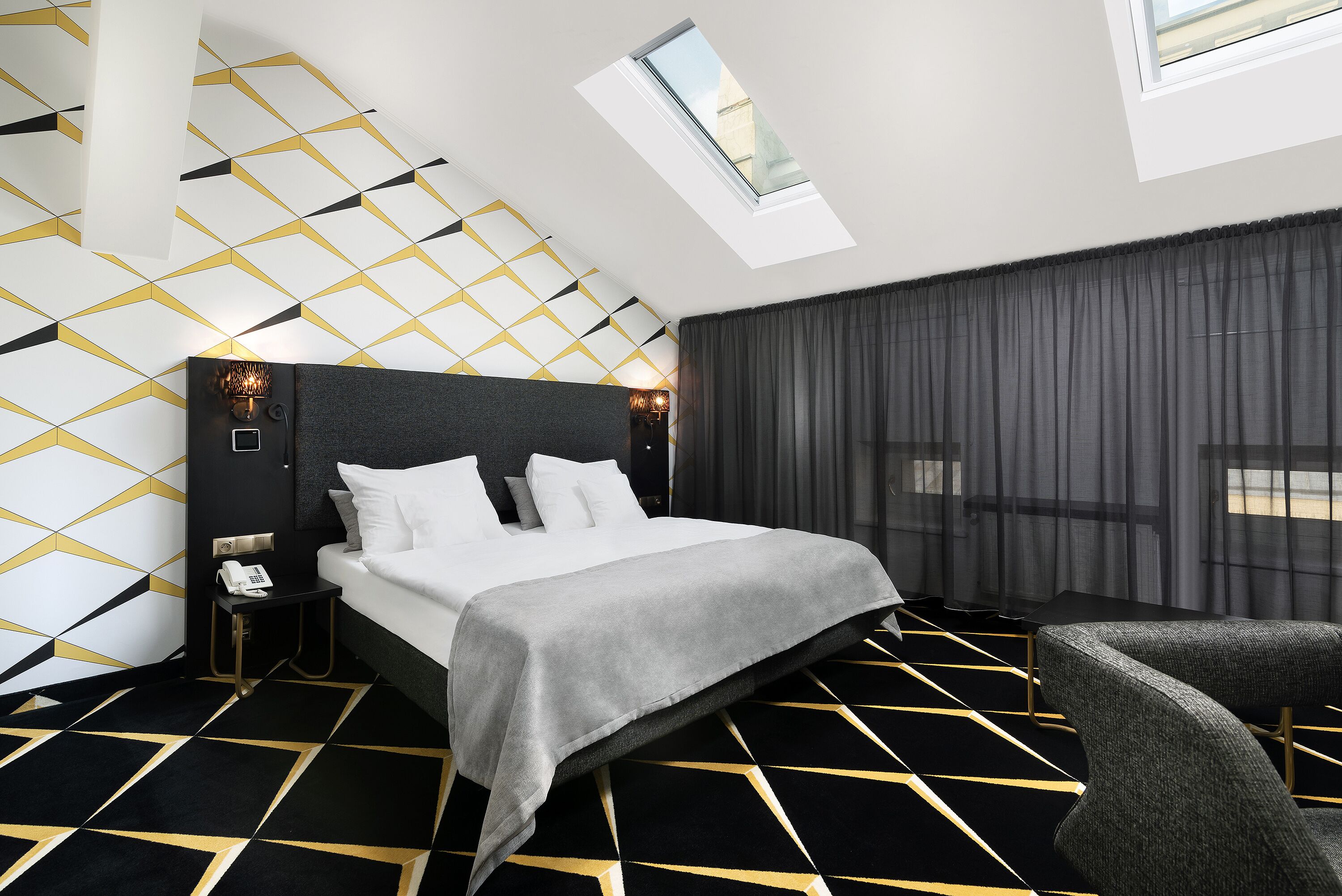 A modern hotel room with a geometric patterned floor, a large bed with a grey bedspread, a bedside table with a phone, and a window with a view of the city.