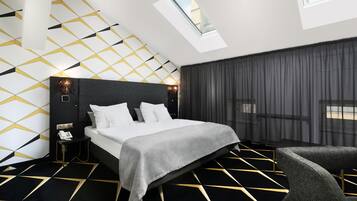 A modern hotel room with a geometric patterned floor, a large bed with a grey bedspread, a bedside table with a phone, and a window with a view of the city.