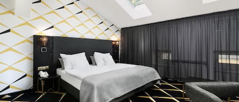 Una habitación de hotel moderna con piso de diseño geométrico, una cama grande con colcha gris, una mesita de noche con teléfono y una ventana con vista a la ciudad.