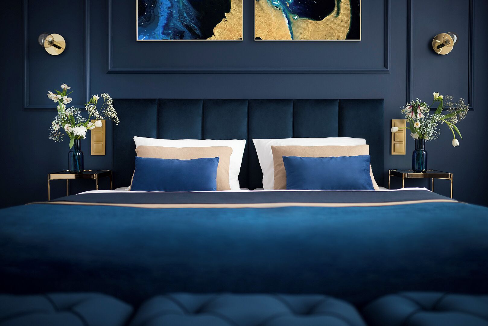 A bedroom with a dark blue color scheme, a tufted headboard, two bedside tables with vases, and a large abstract painting above the bed.