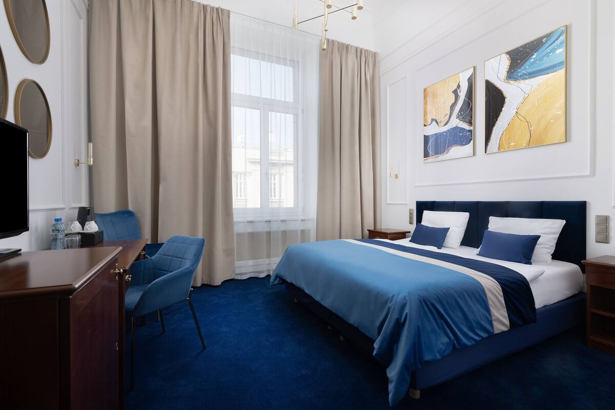 A hotel room with a large bed, a desk, a chair, and artwork on the wall.