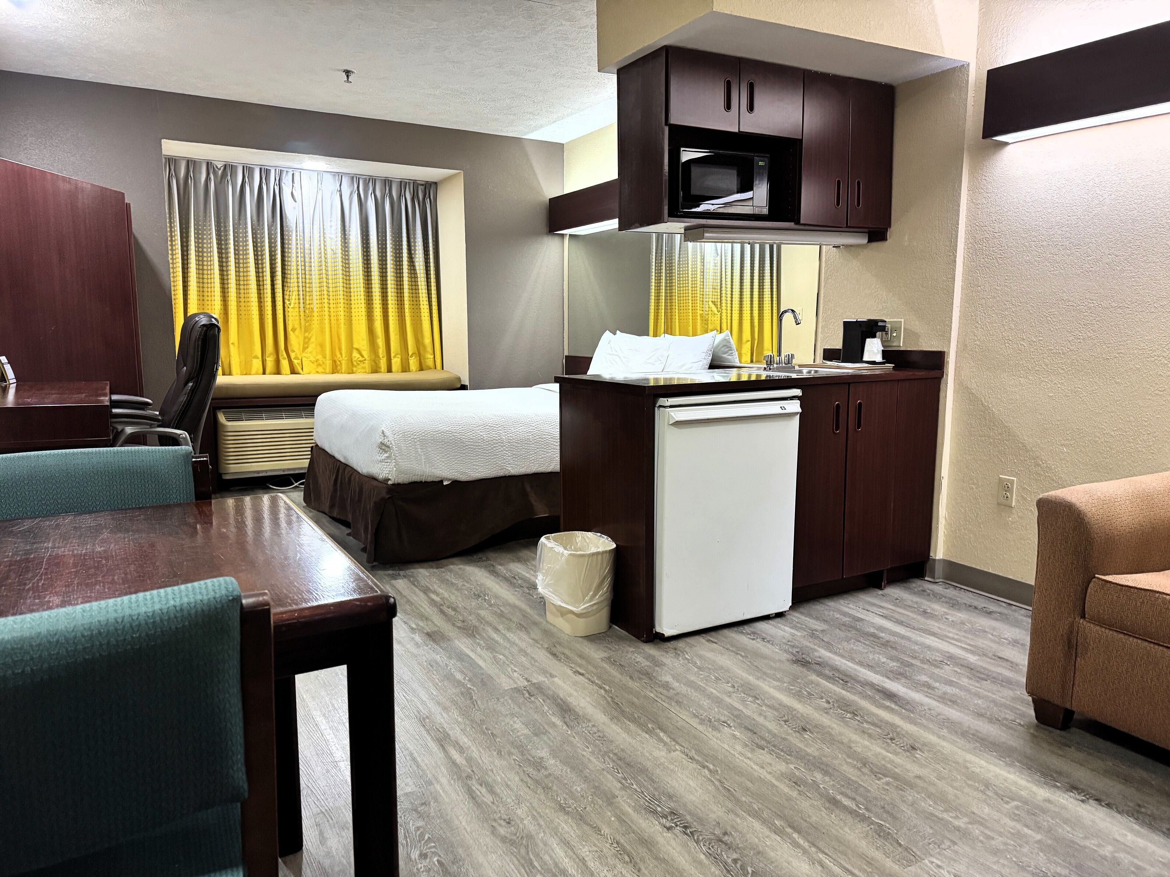 Habitación de hotel con cama, escritorio, silla, kitchenette pequeña y ventana con cortinas.