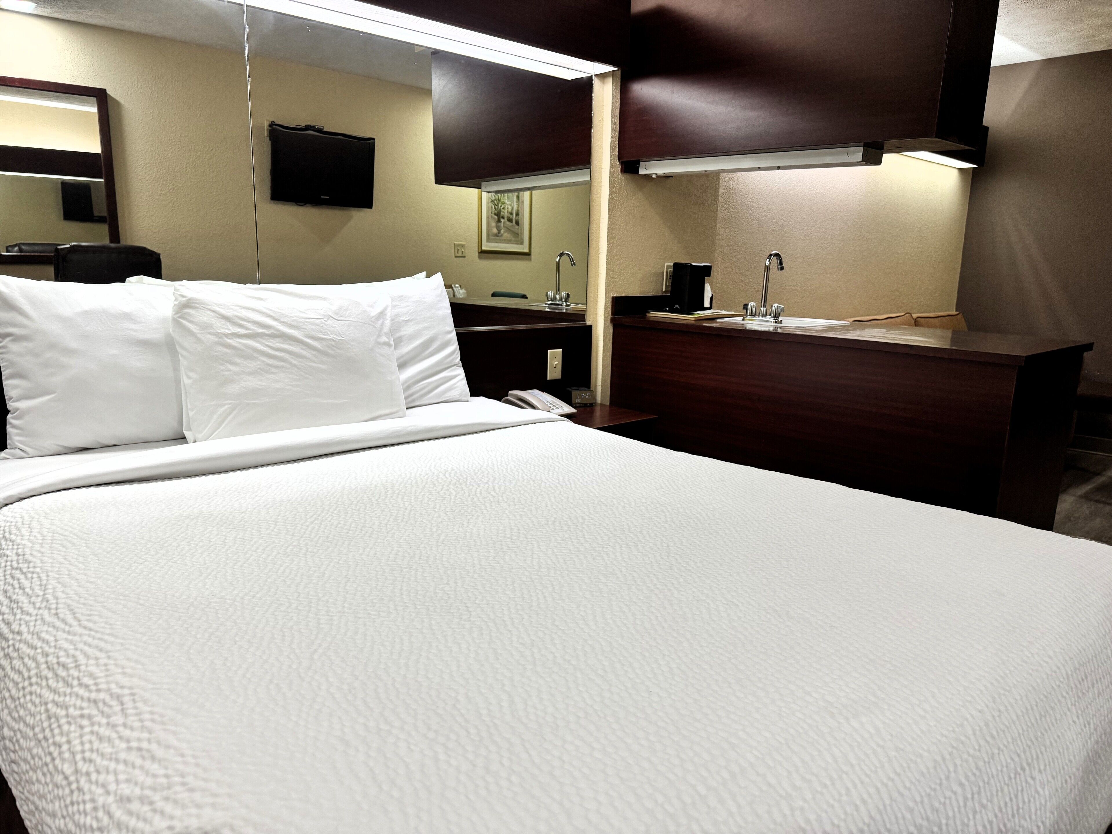 Een hotelkamer met een groot bed, nachtkastje, spiegel, televisie, bureau met stoel, en een badkamer met wastafel en spiegel.
