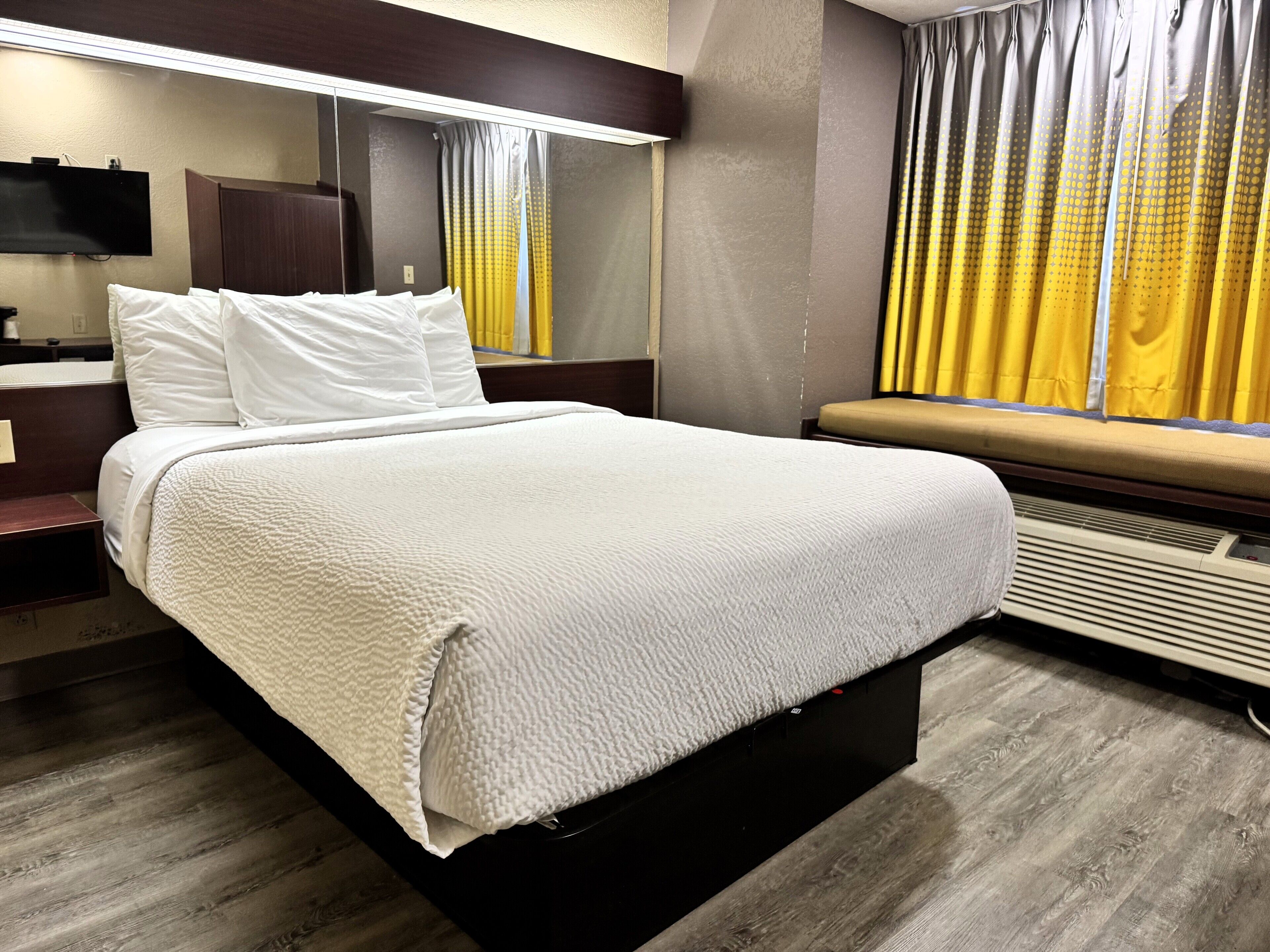A hotel room with a large bed, a bedside table, a flat-screen TV, a window with yellow curtains, and a built-in air conditioning unit.