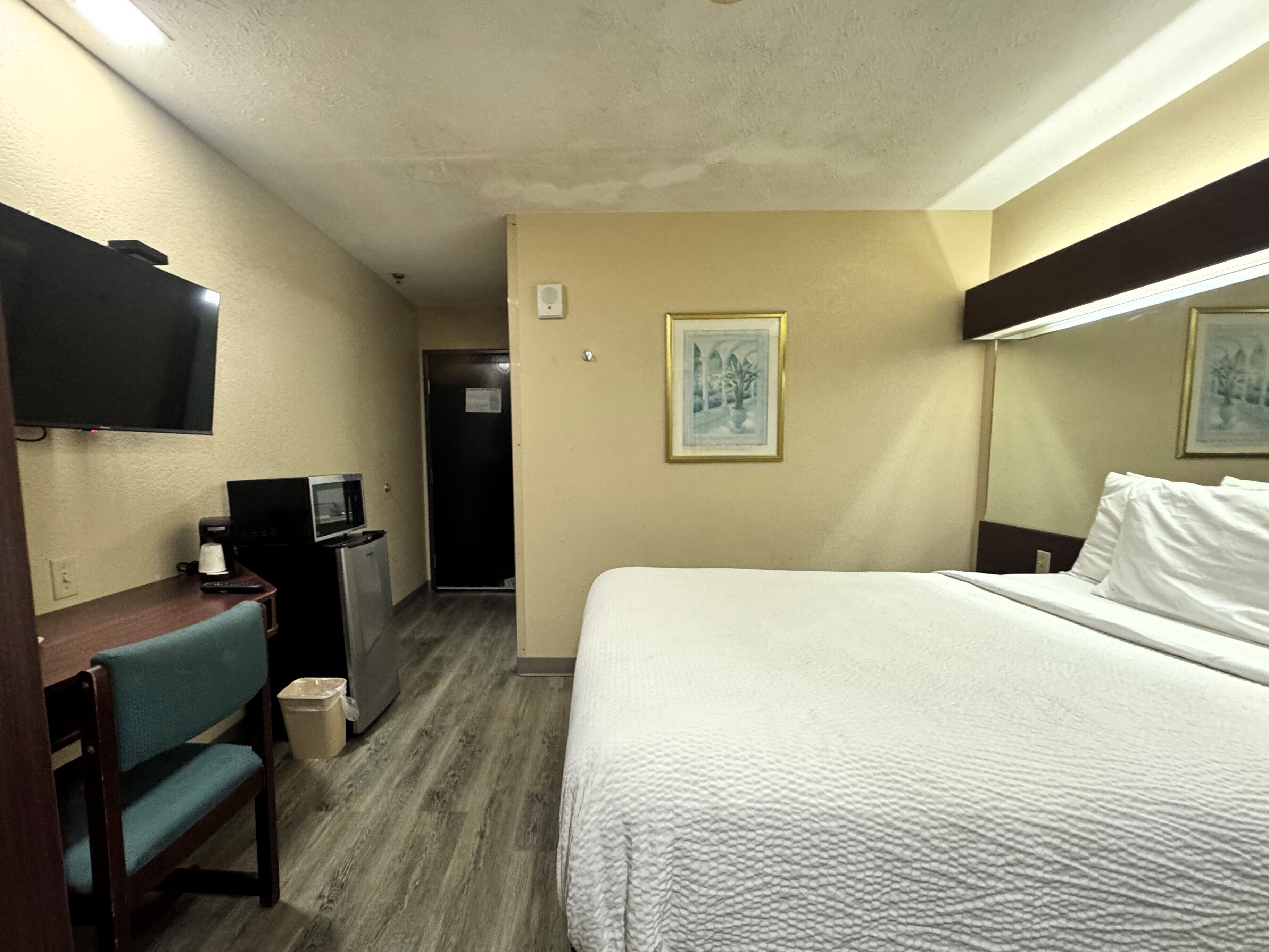 Una habitación de hotel con cama, escritorio con silla, televisión, refrigerador y un cuadro en la pared.