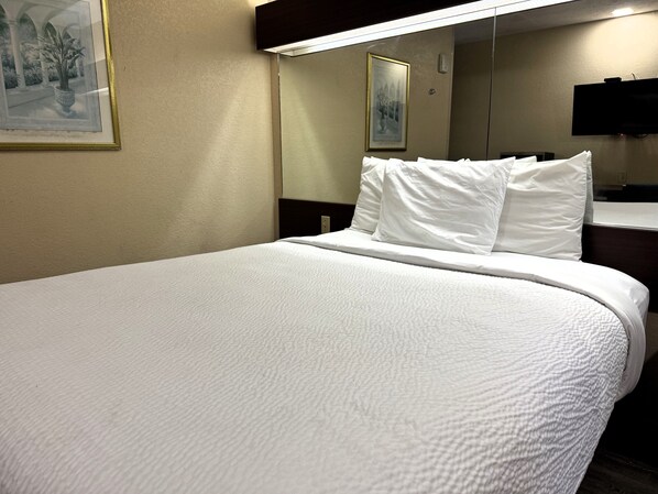 Une chambre d’hôtel avec un grand lit, du linge de lit blanc, un téléviseur à écran plat fixé au mur et un tableau encadré au-dessus du lit.