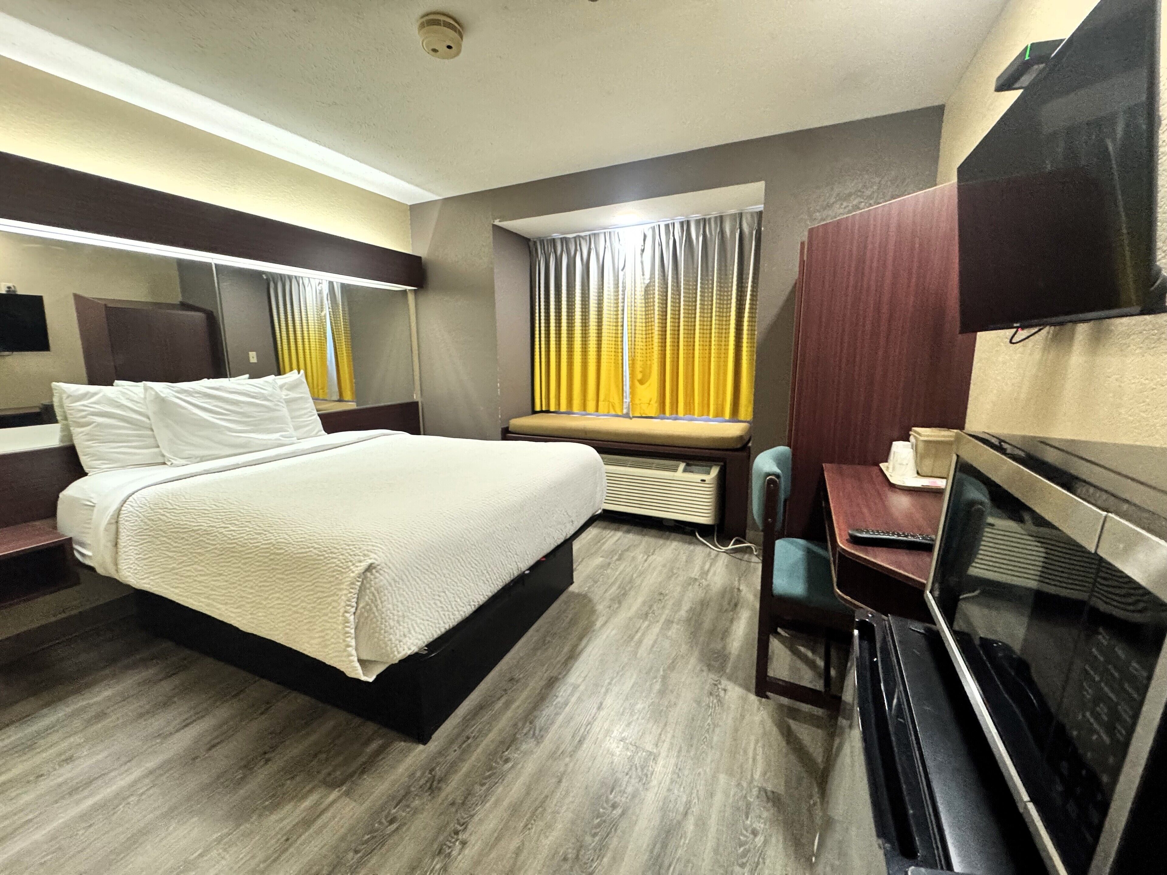 Habitación de hotel con cama, escritorio, silla, televisor y ventana con cortinas.