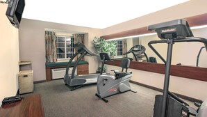 A hotel room with a treadmill, a stationary bike, a mirror, a window with curtains, a plant, and a small trash bin. - Microtel Inn & Suites by Wyndham Meridian (Meridian)