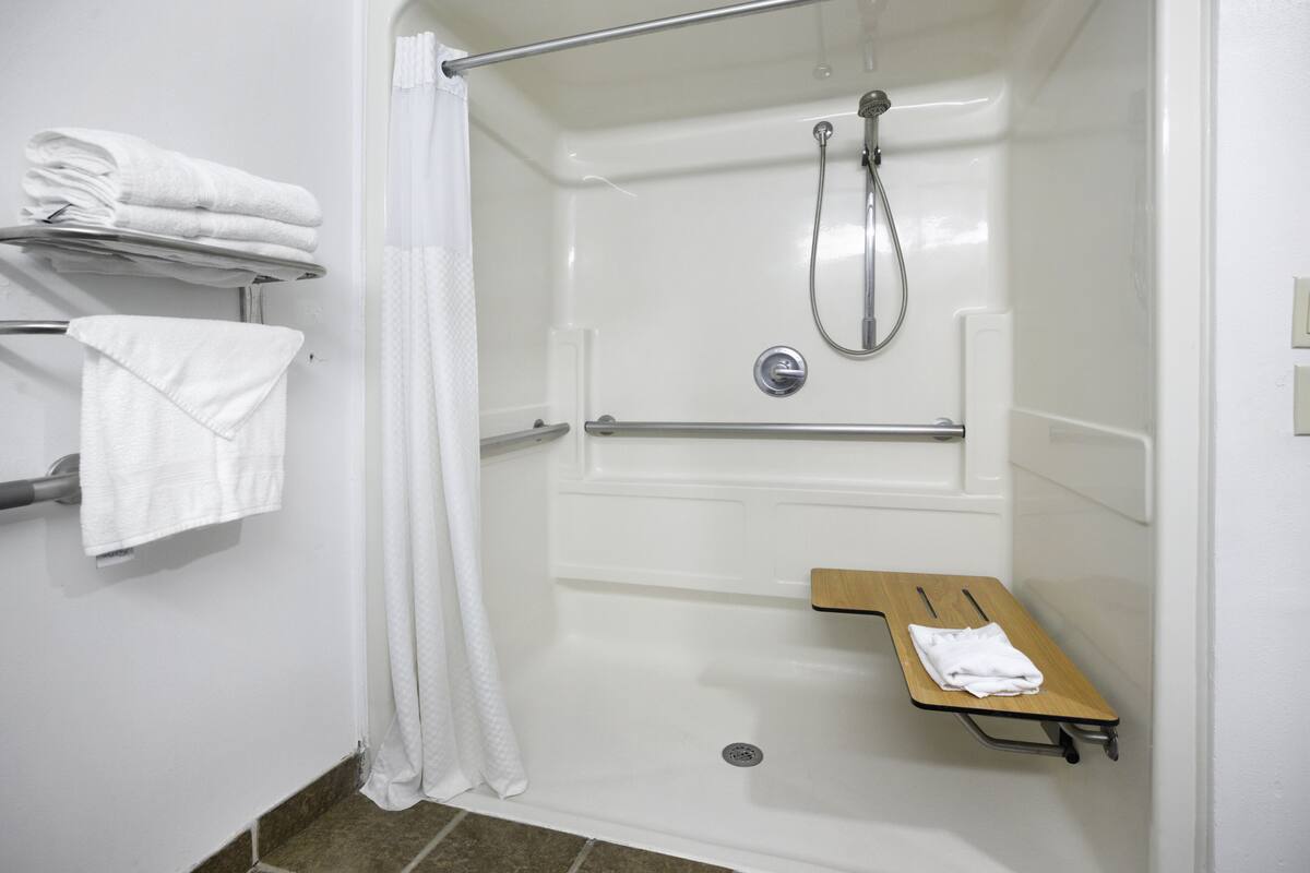 A white shower area with a showerhead, a grab bar, and a small wooden shelf with a towel.