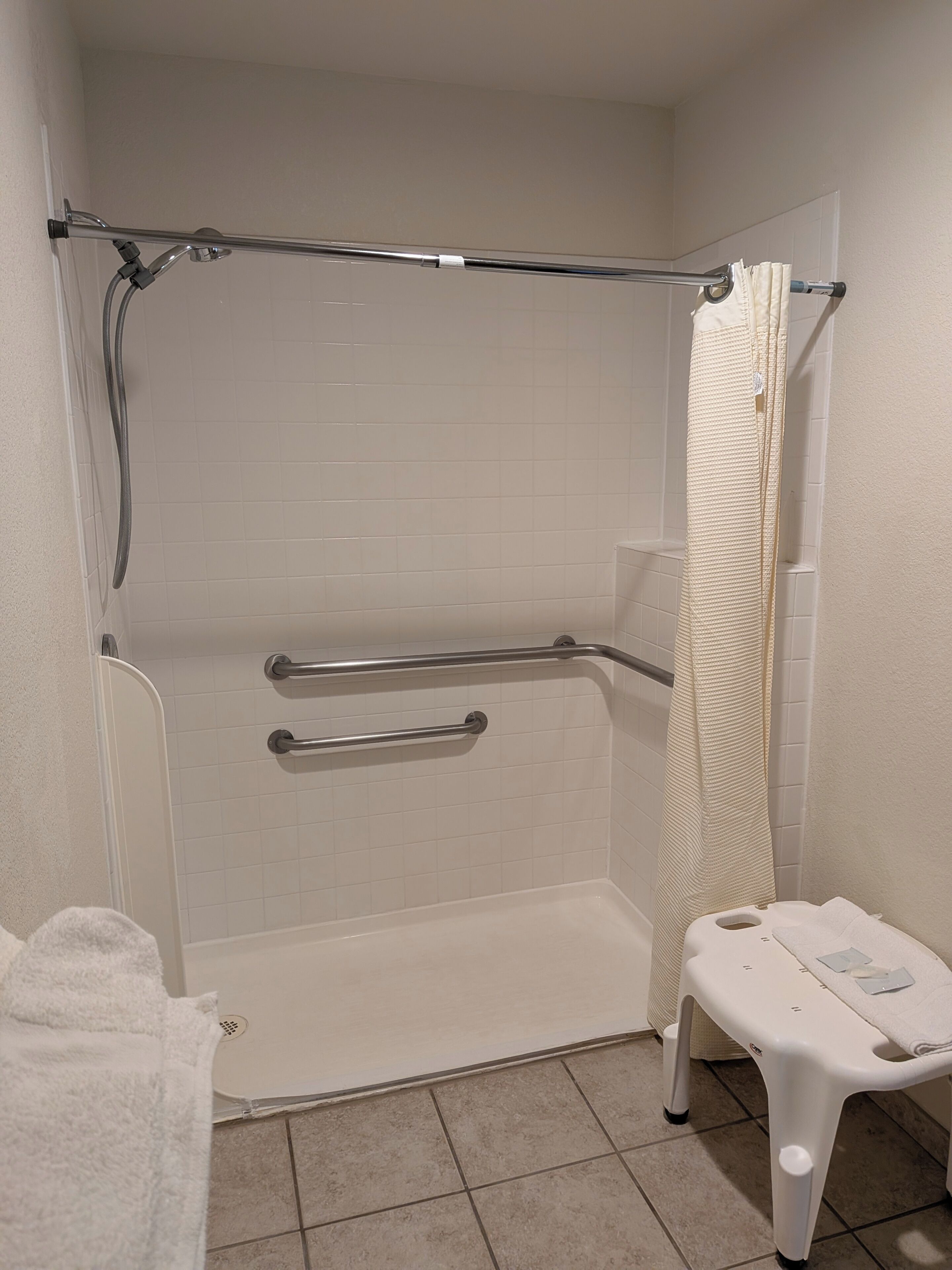 A bathroom with a shower, grab bars, a white towel, and a small white stool.