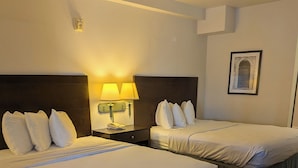A hotel room with two beds, a nightstand with a lamp, a framed picture on the wall, and a patterned carpet. - Catalina Inn (Tucson)