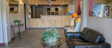 A spacious living area with a leather sofa, a glass coffee table with plants, a kitchen in the background, and a tiled floor.