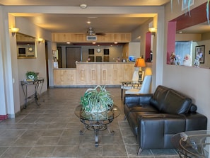 A spacious living area with a leather sofa, a glass coffee table with plants, a kitchen in the background, and a tiled floor.