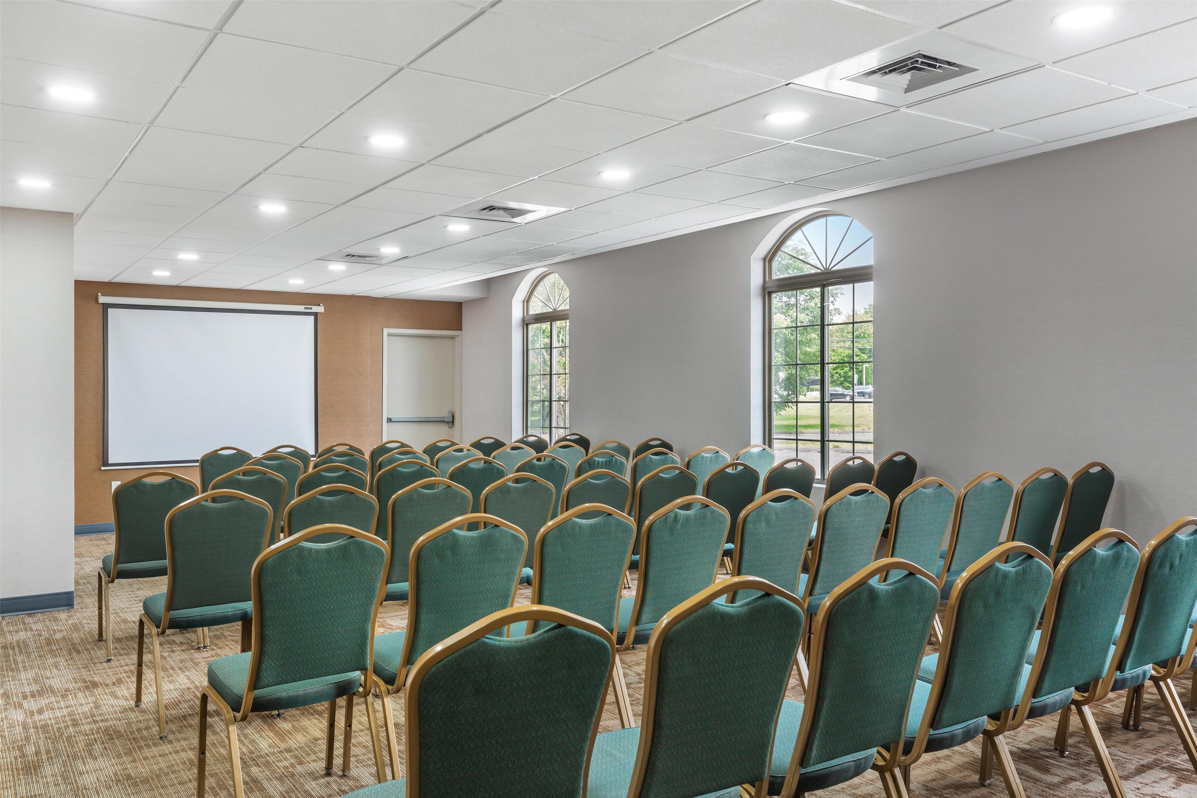 A conference room with rows of teal chairs, a white projection screen, and large arched windows.